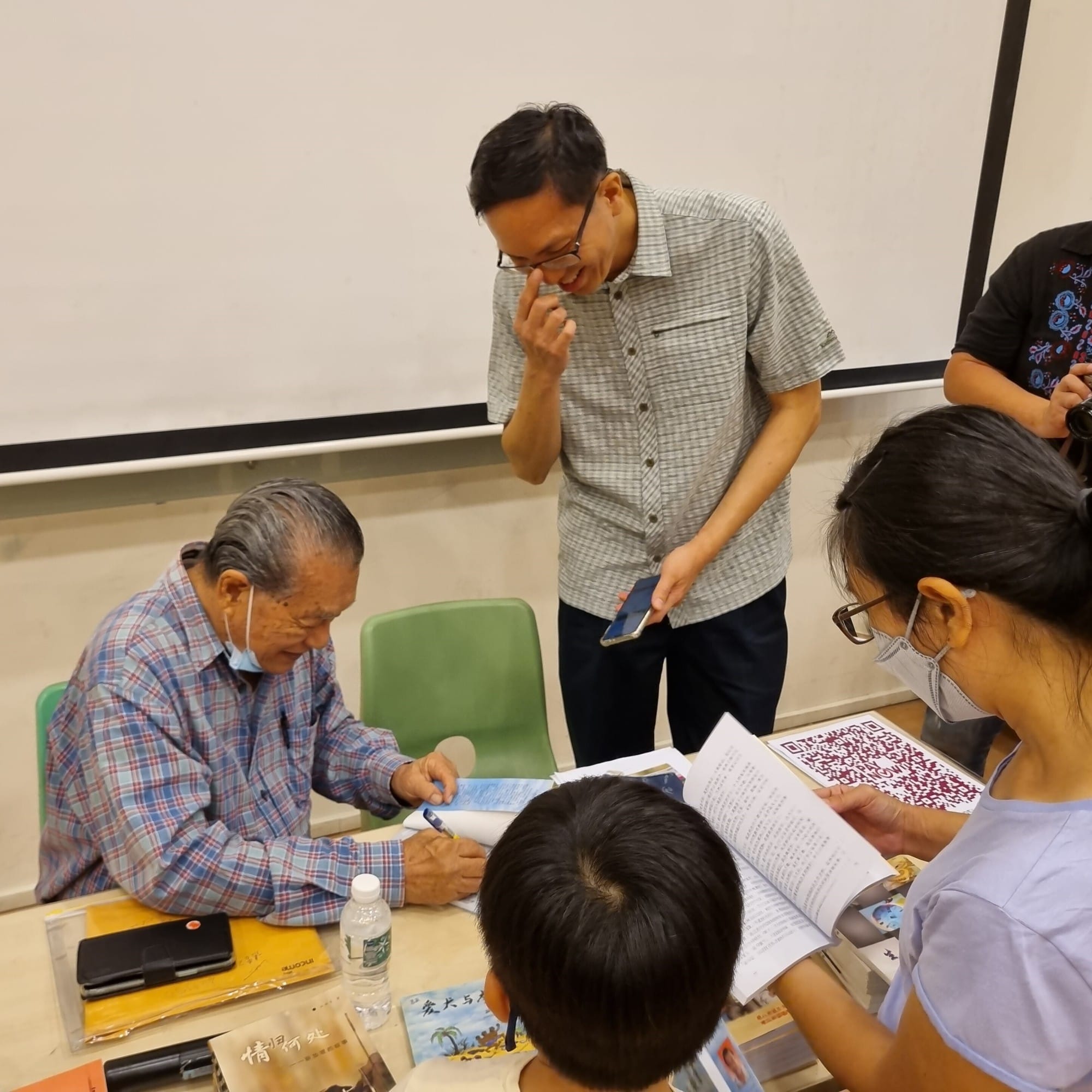 An elderly man signs a book at a table while several people stand around watching in a classroom setting.
